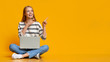 Cheerful teenage girl sitting with laptop, pointing at yellow background