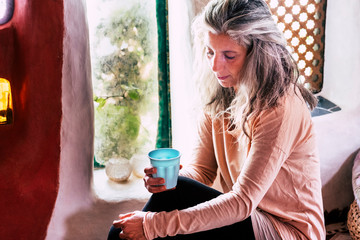 Sad and lonely adult woman with alternative long gy hair and blue cup of tea alone at home near the window - depresion and sadness illness concept