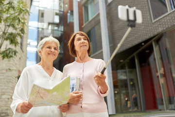 Wall Mural - tourism, travel and friendship concept - happy senior women with map and city guide taking picture by smartphone on selfie stick on street in tallinn