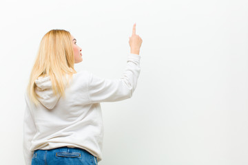 young blonde woman standing and pointing to object on copy space, rear view against white wall