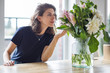 © papa - Portrait of a young beautiful woman sitting at a table with a bouquet of flowers in a vase in a bright room.