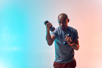 Great workout. Muscular young african man exercising with dumbbells while standing against colorful background