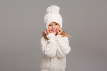 Winter Clothes. Portrait Of A Little Curly-haired Girl In A Knitted White Winter Hat. Little Blonde Girl In White Knitted Hat And Sweater Smiling Light Background Isolate, Space For Text