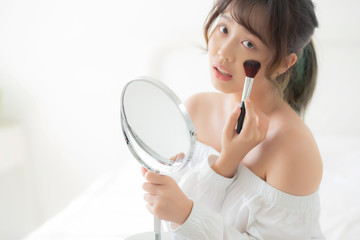 Wall Mural - Beauty portrait young asian woman smile with face looking mirror applying makeup with brush cheek in the bedroom, beautiful of girl holding blusher, skin care and cosmetic concept.