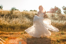 Bride On A Summer Field In White Wedding Dress Rolling And Dancing In Sunset Light. Sun Beams Seen Through Transparent Dress Skirt Fabric. Rustic Or Boho Outdoor Wedding Concept. Selective Soft Focus.