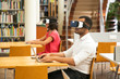 © Mangostar - Adult students using VR glasses in public library. Man and woman wearing virtual reality glasses, sitting at desks and using laptop or tablet in computer class. Innovation concept