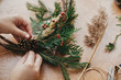 Making rustic christmas wreath. Hands holding pine cones, berries, fir branches, thread, scissors on wooden table.  Authentic rural wreath. Christmas wreath workshop