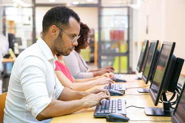 Wall Mural - Extremely focused male student taking online test in computer class. Line of man and women in casual sitting at table, using desktops, typing, looking at monitor. Education center concept