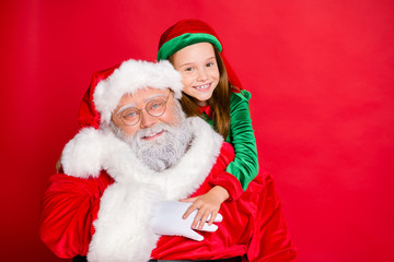 Sticker - Close up photo of charming little elf helper in green hat headwear hugging piggyback her funny santa claus wearing eyewear eyeglasses isolated over red background