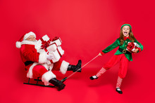 Full Size Photo Of Funny Santa Claus With Eyeglasses Eyewear Spectacles And Cute Elf In Green Hat Headwear Holding Sledges Carry Sack Bag With Gifts Isolated Over Red Background