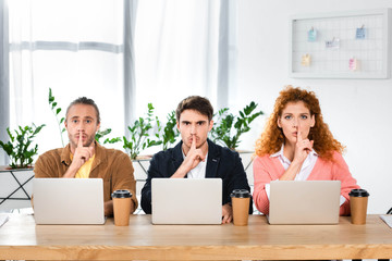 three friends sitting at table and showing shh gestures in office