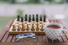 Close Up Photo Of Glasses, White Cup Of Black Tea And Chess On Wooden Table. Chess Pieces Ready For The Game.Cozy Evenings, Home Atmosphere. Concept Of Comfort And Coziness. Autumn Mood. Side View.
