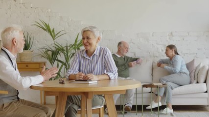 Wall Mural - Tracking shot of elderly man and woman sitting at table and enjoying talk, another senior couple playing cards in background sitting on sofa in common room of assisted living home