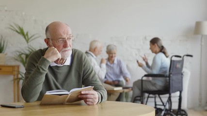 Wall Mural - Rack focus of three elderly people, two women including disabled one and man, playing cards in nursing home. Intelligent senior man in eyeglasses reading book and thinking sitting at table