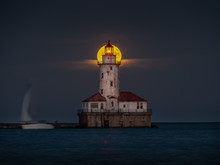 A Beautiful Night Shot Of The Harvest Full Moon As It Aligns With The Center Top Of The Abandoned Historic Light House Tower Along Lake Michigan In Chicago As Sailboat Passes By On The Dark Water.