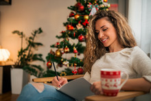 Portrait Of A Smiling Young Woman Writing Christmas Cards In Front Of Tree.