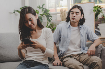 young man look at her girlfriend from behind who ignore and focused into mobile phone sitting on sofa in living room at home together, upset couple, divorce, family issues and relationship concept