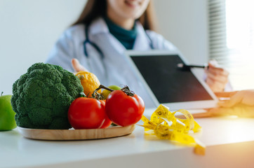 Wall Mural - focus fresh vegetables and fruits with nutritionist female doctor using tablet and talking about diet plan with patient on desk at office hospital, food science, healthy food and dieting concept