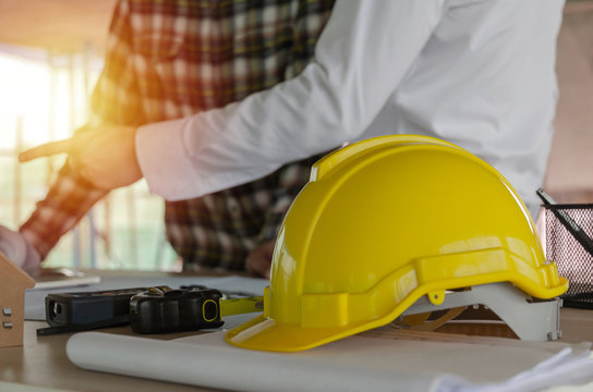 yellow safety helmet on workplace desk with construction worker team engineer or inspector checking new project contract at construction site background, engineering, construction, contractor concept