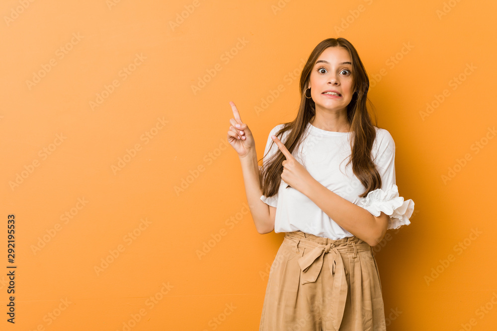 Young caucasian woman shocked pointing with index fingers to a copy space. - obrazy, fototapety, plakaty 