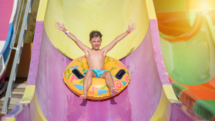 Happy boy on water slide in a swimming pool having fun during summer vacation in beautiful aqua park. Boy slithering down water slide.