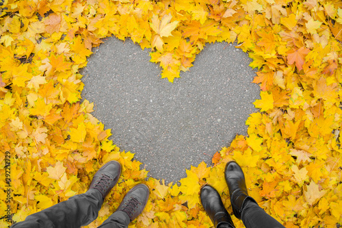 Young couple\'s legs standing beside each other. Heart shape created from yellow, orange, lush maple leaves. Asphalt surface in middle. Empty place for positive text, quote or sayings. Top view.
