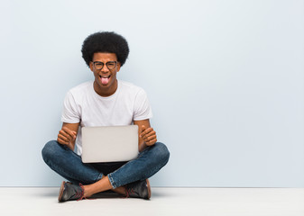 Wall Mural - Young black man sitting on the floor with a laptop funnny and friendly showing tongue