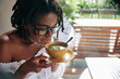 © DragonImages - African young woman with beautiful hairstyle and in eyeglasses holding bowl with soup and she is going to taste it