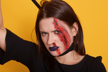 Horizontal Shot Of Young Female With Dangerous Facial Expression Andbloody Wound On Her Face, Lady Choking Herself With Piece Of Cloth, Having Dark Hair, Wearibg Black Dress. Halloween Holiday Concept