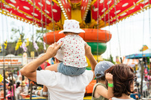 Child Sitting On Her Father's Shoulders In Front Of A Colorful Carousel At A Carnival. Girl And Boy With Their Parents At The Vintage Amusement Park Rides. Wearing A Cute Ladybug Outfit And A Sun Hat.