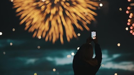 Happy woman shoots beautiful salute in the night sky of a megalopolis on a smartphone. Silhouette of people watching explosive and colorful fireworks in the evening sky. Holiday event and celebration.