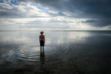 Tropical Silhouette Of Man In Straw Hat Standing In Calm Shallow Waters With Ripples Emanating On The Surface