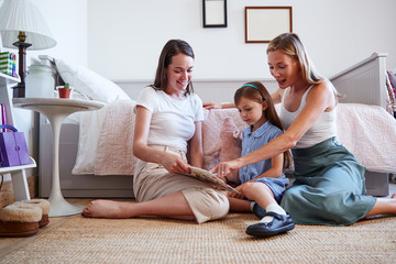 Same Sex Female Couple Sitting On Bedroom Floor Reading Book With Daughter At Home Together