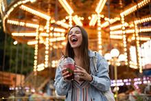 Outdoor Portrait Of Joyful Young Pretty Brunette Female In Casual Clothes Posing Over Amusement Park With Closed Eyes And Broad Smile, Holding Cup Of Lemonade In Hands