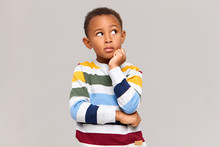 Studio Picture Of Cute Dark Skinned Nine Year Old Boy Keeping Hand Under His Chin And Looking Up With Thoughtful Pensive Facial Expression, Having Curious Look, Trying To Remember Something