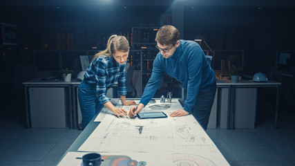 In the Dark Industrial Design Engineering Facility Male and Female Engineers Talk and Work on a Blueprints Using Conference Table. On the Desktop Drawings, Drafts and Electric Engine Components, Parts