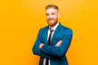 © kues1 - young red head businessman smiling to camera with crossed arms and a happy, confident, satisfied expression, lateral view against orange background