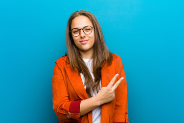 Wall Mural - young woman feeling happy, positive and successful, with hand making v shape over chest, showing victory or peace against blue background