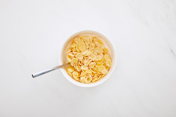 top view of bowl of cornflakes with spoon on white marble surface