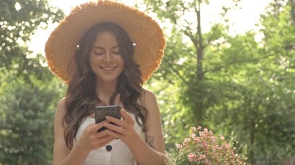 Canvas Print - Happy pretty young brunette woman in straw hat smiling and using smartphone while walking in the park