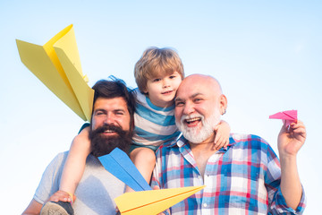 Wall Mural - Multi generation. Portrait of happy father giving son piggyback ride on his shoulders and looking up. Happy child playing with toy paper airplane against summer sky background.