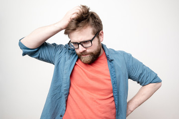 Young bearded man with glasses is in doubt, he is confused, looks down thoughtfully and scratches his head, on a white background.