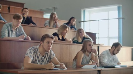 modern students, men and women, sit at desks in a large classroom and write a lecture notes.