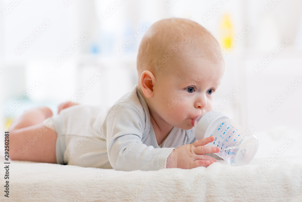 Pretty baby drinks water from bottle lying on bed in nursery room. - obrazy, fototapety, plakaty 