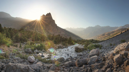 Fototapete - Mountain valley. Bright mountains nature landscape at sunrise. Mountain river and vivid sun rays behind mount peak. Scenic highland in warm sunlight