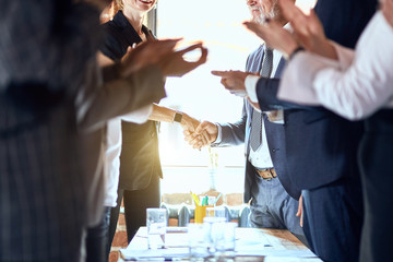 Wall Mural - Group of businessmen at table in office. Focuced on shaking hands. Others applaud.