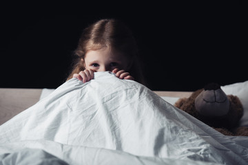 Wall Mural - scared child hiding under blanket while sitting on bedding near teddy bear isolated on black