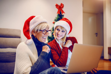 Wall Mural - Happy senior woman and her daughter sitting on floor in living room and using laptop for christmas shopping. Both having santa hats on heads. In background is christmas tree.