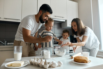 bearded daddy with beautiful woman , their kids holding flour on his palm, adding it to the bowl with dough in the kitchen