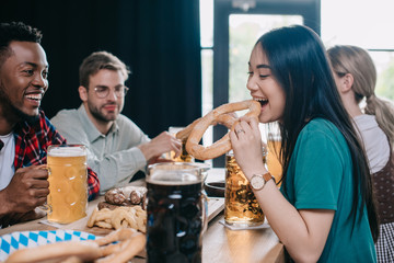 Wall Mural - young asian woman eating pretzel while celebrating octoberfest with multicultural friends in pub